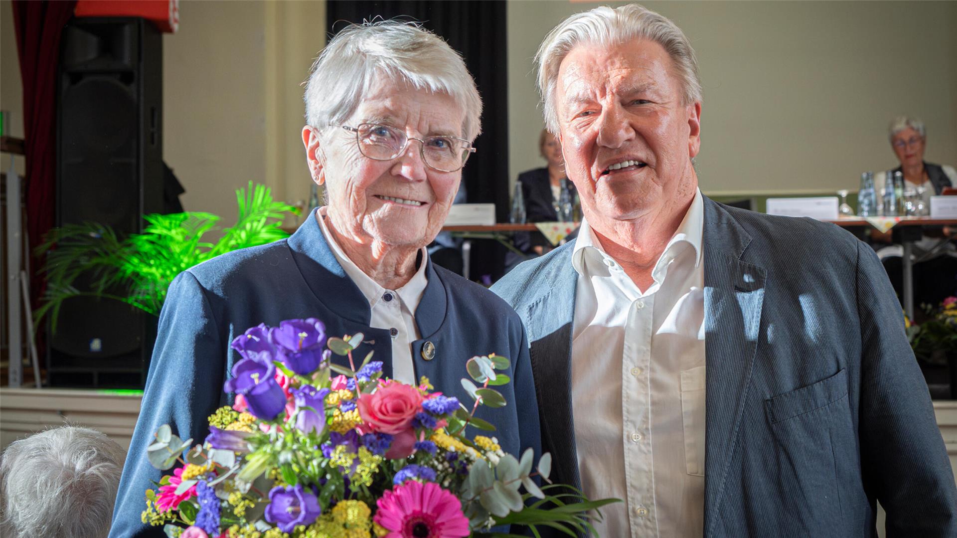 Ältere Frau mit Blumenstrauß steht lächelnd neben einem Mann in hellem Hemd und Sakko. Beide blicken in die Kamera, im Hintergrund läuft eine Veranstaltung.