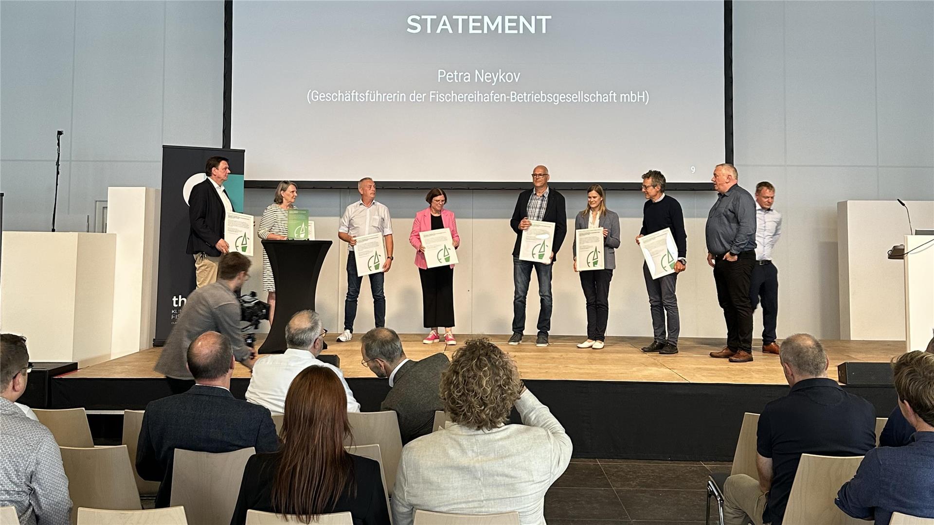 Menschen stehen auf einer Bühne und halten ein Plakat in den Händen. Davor sitzen Menschen auf Stühlen mit dem Rücken zum Betrachter. 