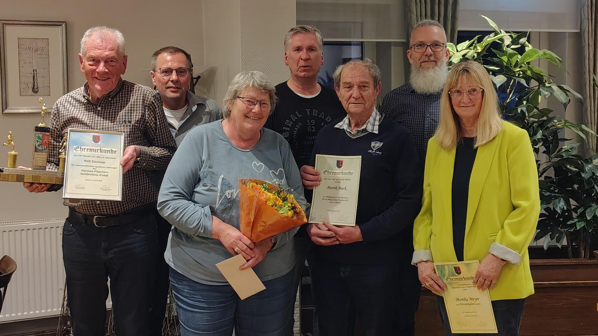 Eine Gruppe von sieben Personen steht zusammen und lächelt in die Kamera. Drei von ihnen halten Ehrenurkunden, eine Frau einen Blumenstrauß. Ein Mann präsentiert einen Pokal. Die Auszeichnung fand offenbar in feierlichem Rahmen statt.