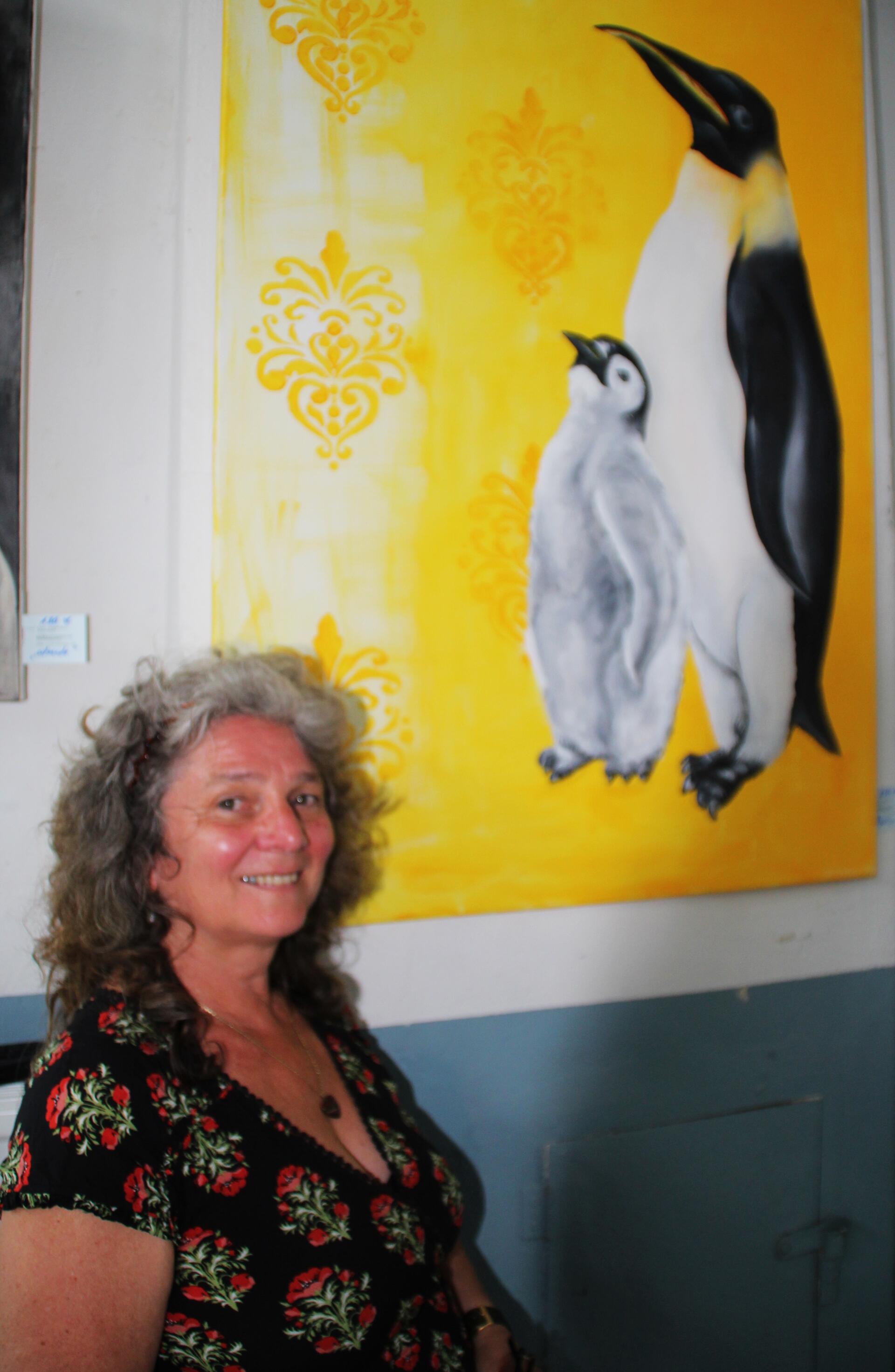 Eine Frau steht vor einem Bild. Abgebildet sind dort auf gelbem Hintergrund zwei Pinguine.