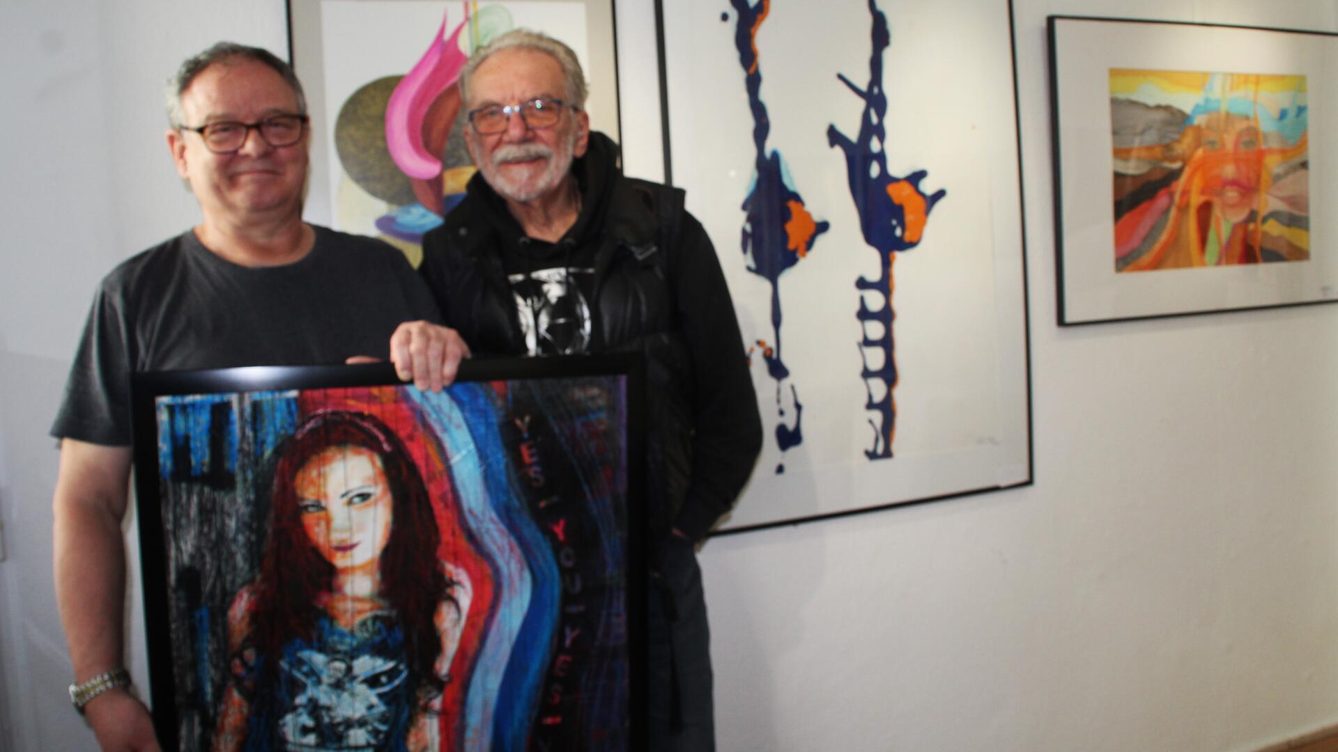 Zwei Männer stehen in einer Galerie nebeneinander und halten ein gerahmtes Gemälde mit einer farbintensiven Frauenfigur. Hinter ihnen hängen mehrere abstrakte Bilder an der weißen Wand.