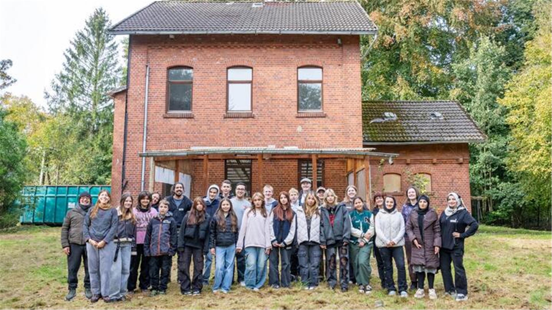 Eine große Gruppe von Jugendlichen und Erwachsenen steht vor einem alten, roten Backsteingebäude mit einem überdachten Eingangsbereich. Die Gruppe besteht aus Menschen verschiedener Altersgruppen und Hintergründe, die lächelnd in die Kamera blicken. 