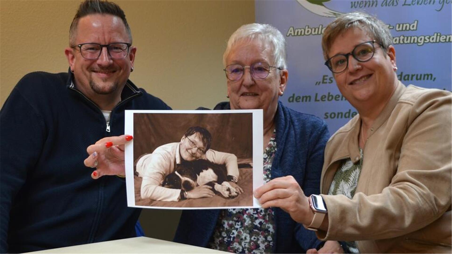 Ein Mann und zwei Frauen. Die Frauen halten ein Foto hoch.