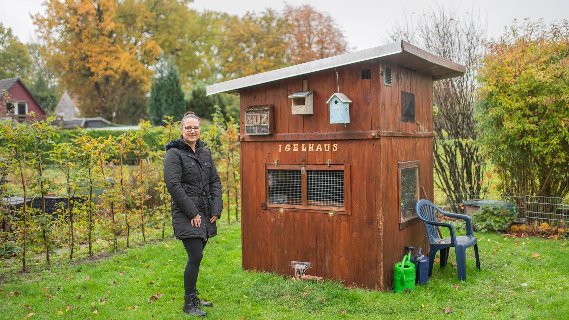 Frau im Garten neben einem Gartenhaus, auf dem "Igelhaus" steht.  
