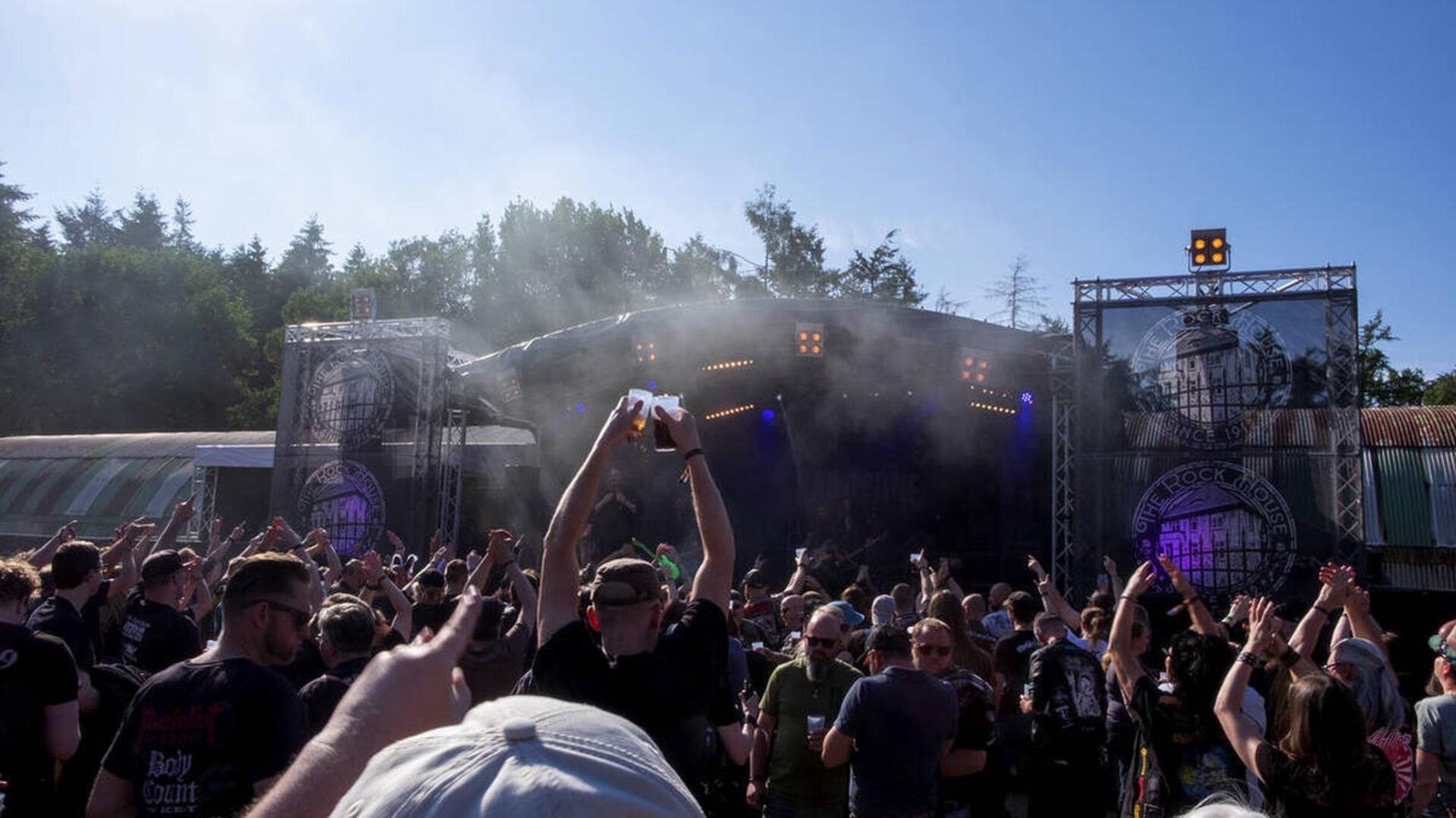 Publikum feiert bei sonnigem Wetter vor einer Open-Air-Bühne auf einem Rockfestival, einige Besucher halten Getränke hoch, im Hintergrund sind Musiker auf der Bühne zu sehen.