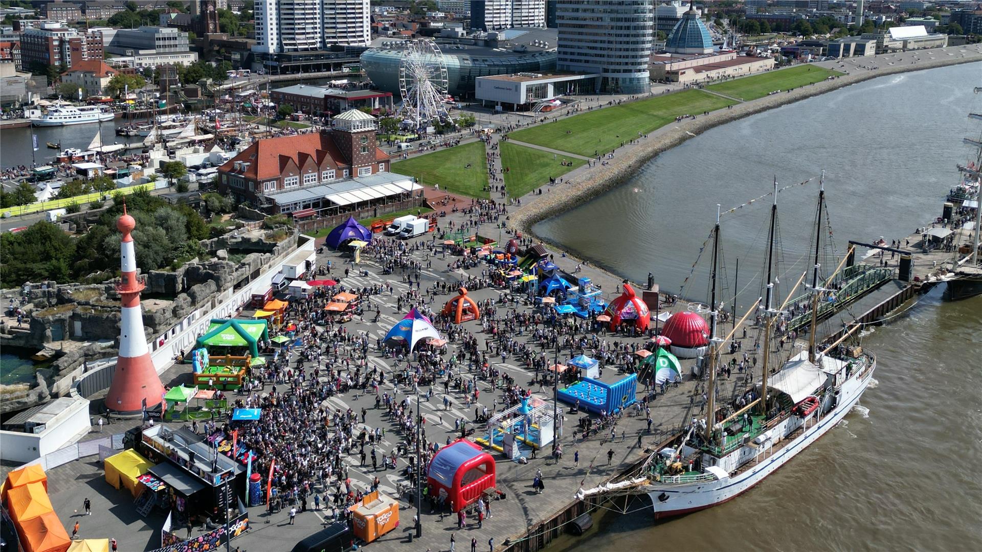 **ALT-Text:**
Luftaufnahme vom Willy-Brandt-Platz in Bremerhaven während eines Großevents mit bunten Pavillons, Aktionsflächen und Menschenmengen am Weserufer.