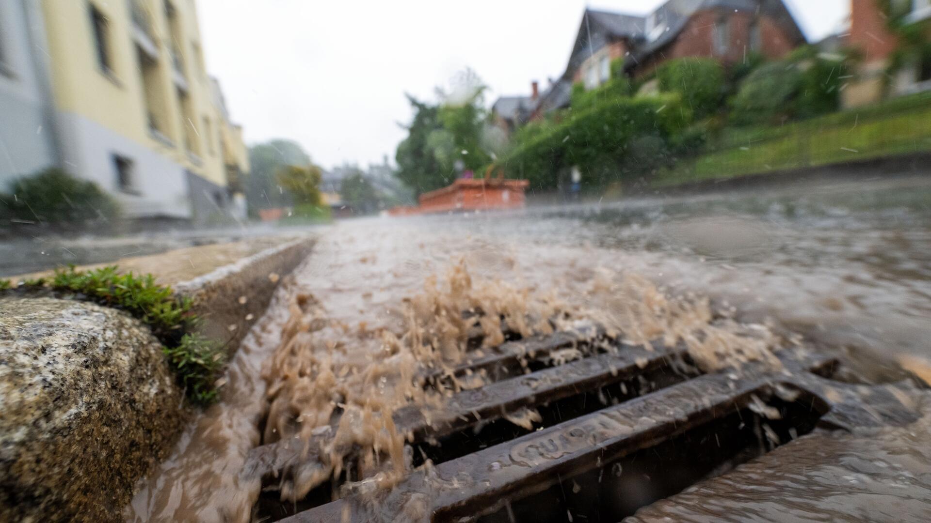 Starkregen überflutet eine städtische Straße; braunes Wasser schießt aus einem überlasteten Gully, im Hintergrund sind Wohnhäuser und Bäume erkennbar.