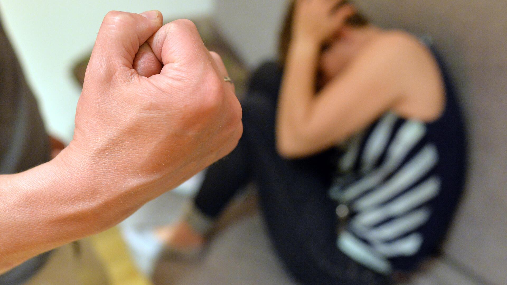 Ein Mann zeigt seine Faust, eine Frau sitzt verängstigt auf dem FJußboden und drückt sich an eine Wand.