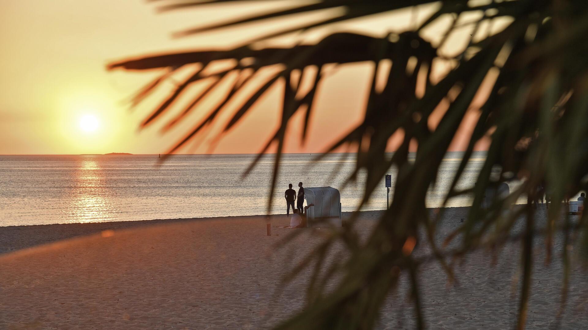 Zwei Menschen stehen neben einem Strandkorb am Strand und beobachten den Sonnenuntergang.