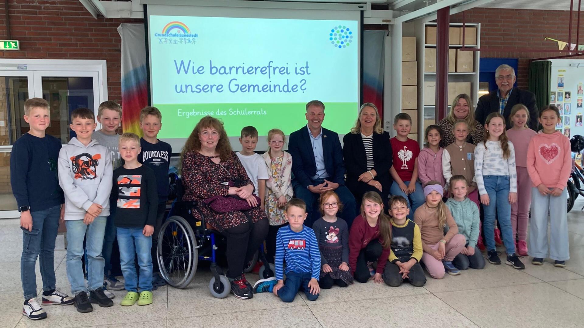 **ALT-Text:**
Gruppenfoto von Grundschulkindern, Lehrkräften, dem Bürgermeister und Mitgliedern des Inklusionsbeirats vor einer Präsentationsfolie mit der Aufschrift „Wie barrierefrei ist unsere Gemeinde? – Ergebnisse des Schülerrats“.