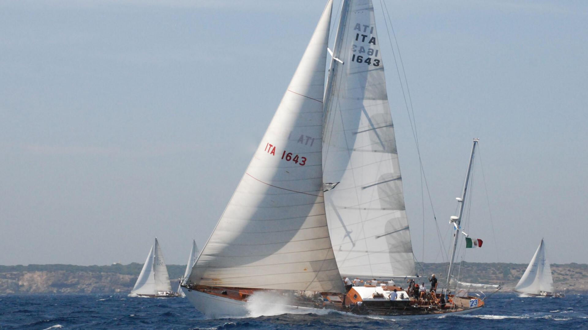 Die italienische Segelyacht „Corsaro II“ unter vollen weißen Segeln auf dem offenen Meer, mit sichtbarer italienischer Flagge am Heck. Weitere Segelschiffe sind im Hintergrund vor einer felsigen Küste zu sehen.