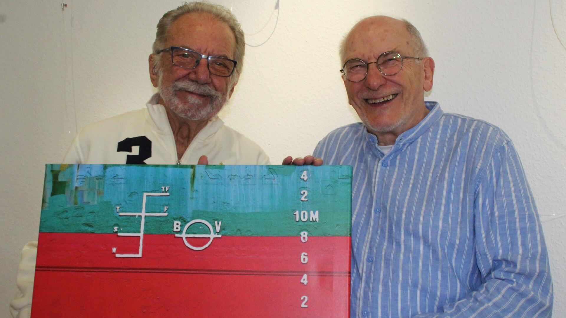 Zwei ältere Männer stehen lächelnd nebeneinander und halten gemeinsam ein farbenfrohes Foto oder Gemälde mit maritimem Motiv, das eine Schiffsbordwand mit Tiefgangmarkierungen und strukturierten Farbflächen zeigt.