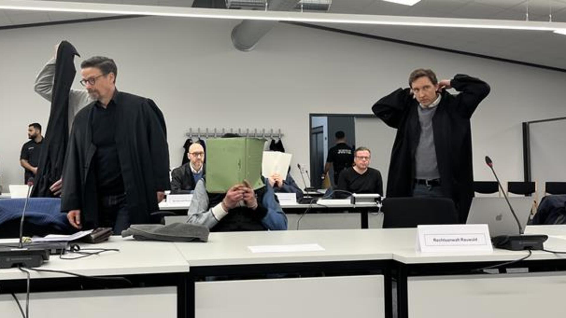 Ein Angeklagter sitzt in einem Gerichtssaal und hält sich eine grüne Mappe vor das Gesicht.