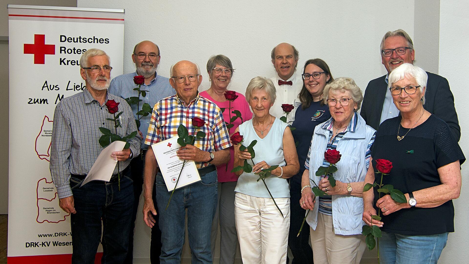 Gruppe von zehn Personen, die beim DRK geehrt wurden, stehen mit roten Rosen und Urkunden in der Hand vor einem Banner des Deutschen Roten Kreuzes.