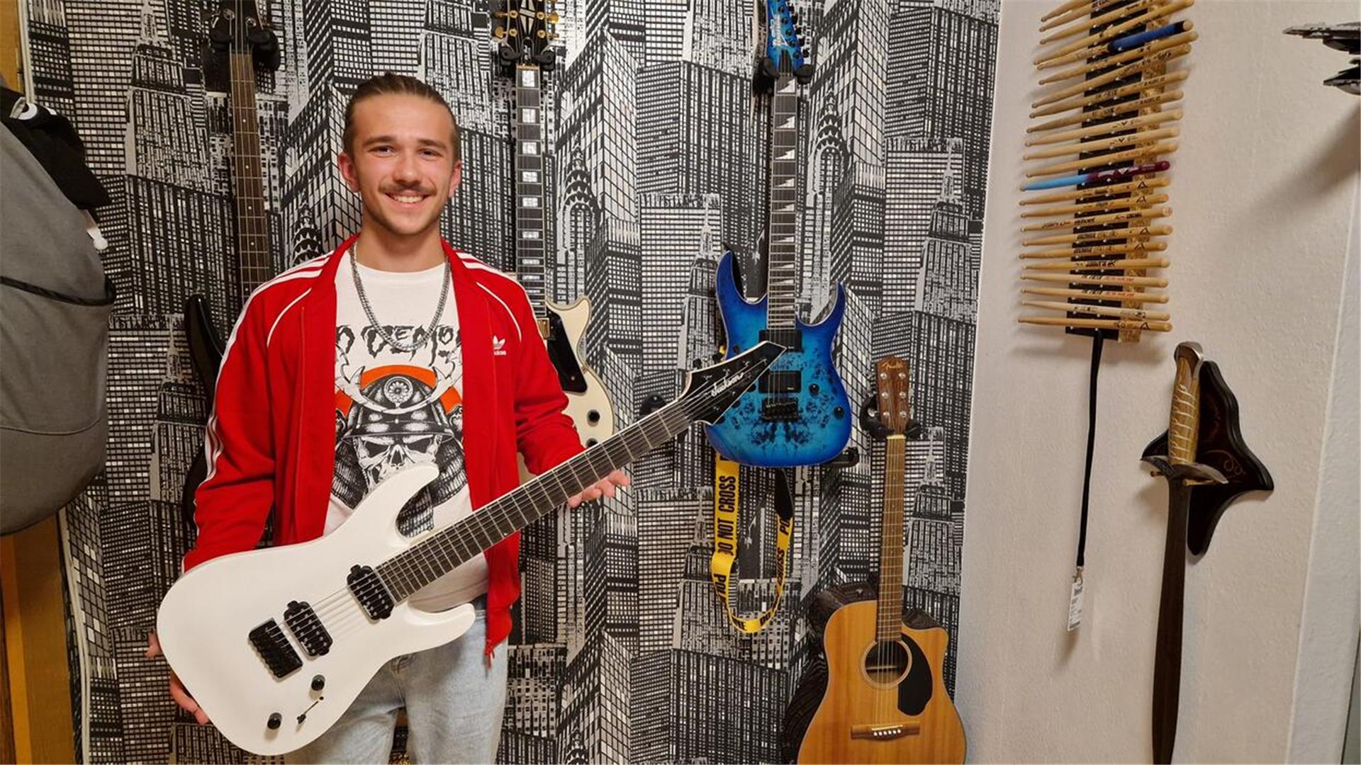 Ein junger Mann steht mit einer Gitarre in der Hand vor einer Wand, an der weitere Gitarren hängen, und lächelt in die Kamera.