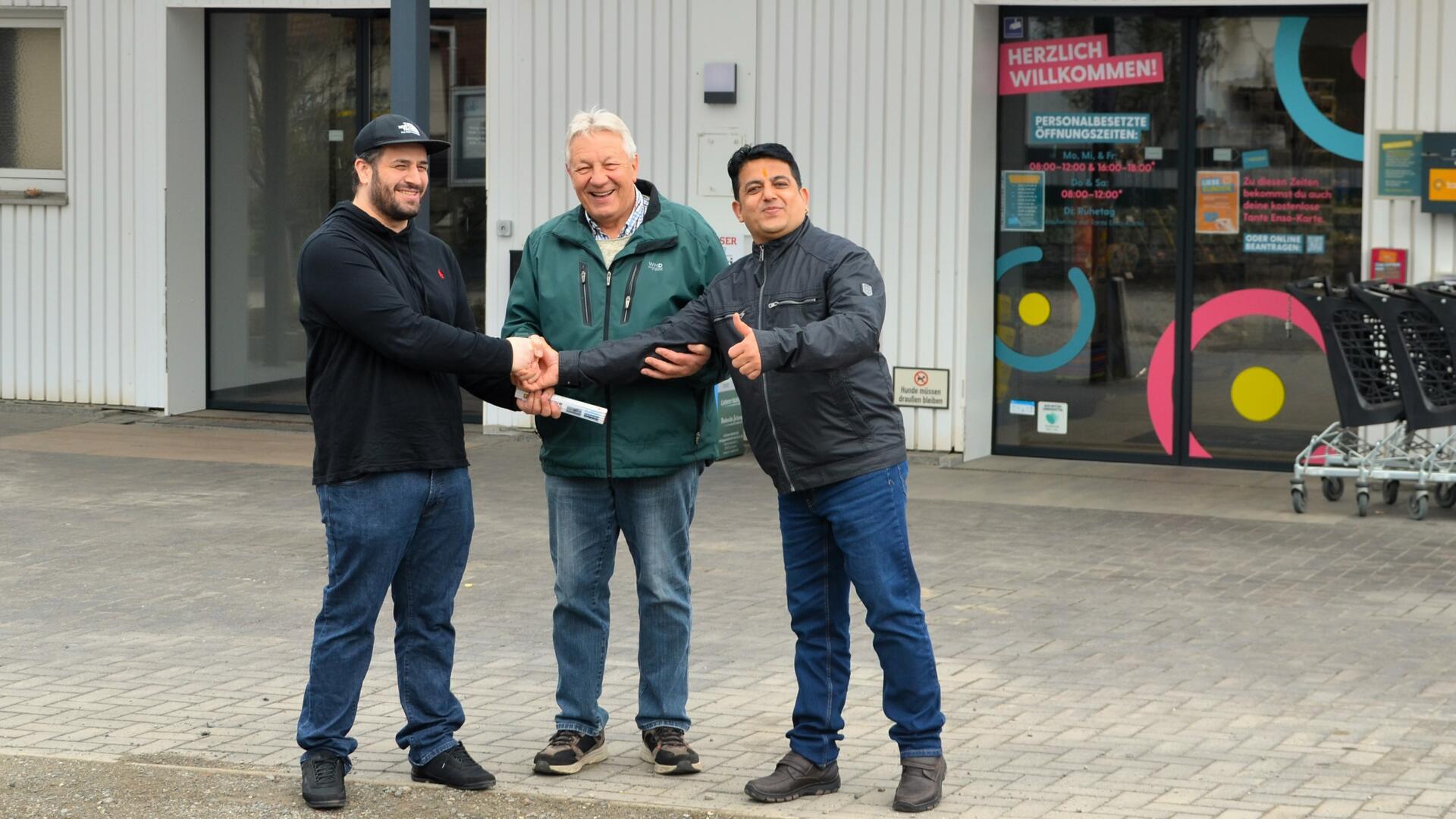 Drei Männer stehen vor einem Ladeneingang, zwei geben sich die Hand, einer zeigt Daumen hoch; alle lächeln, im Hintergrund Einkaufswagen und ein Schild mit Öffnungszeiten.