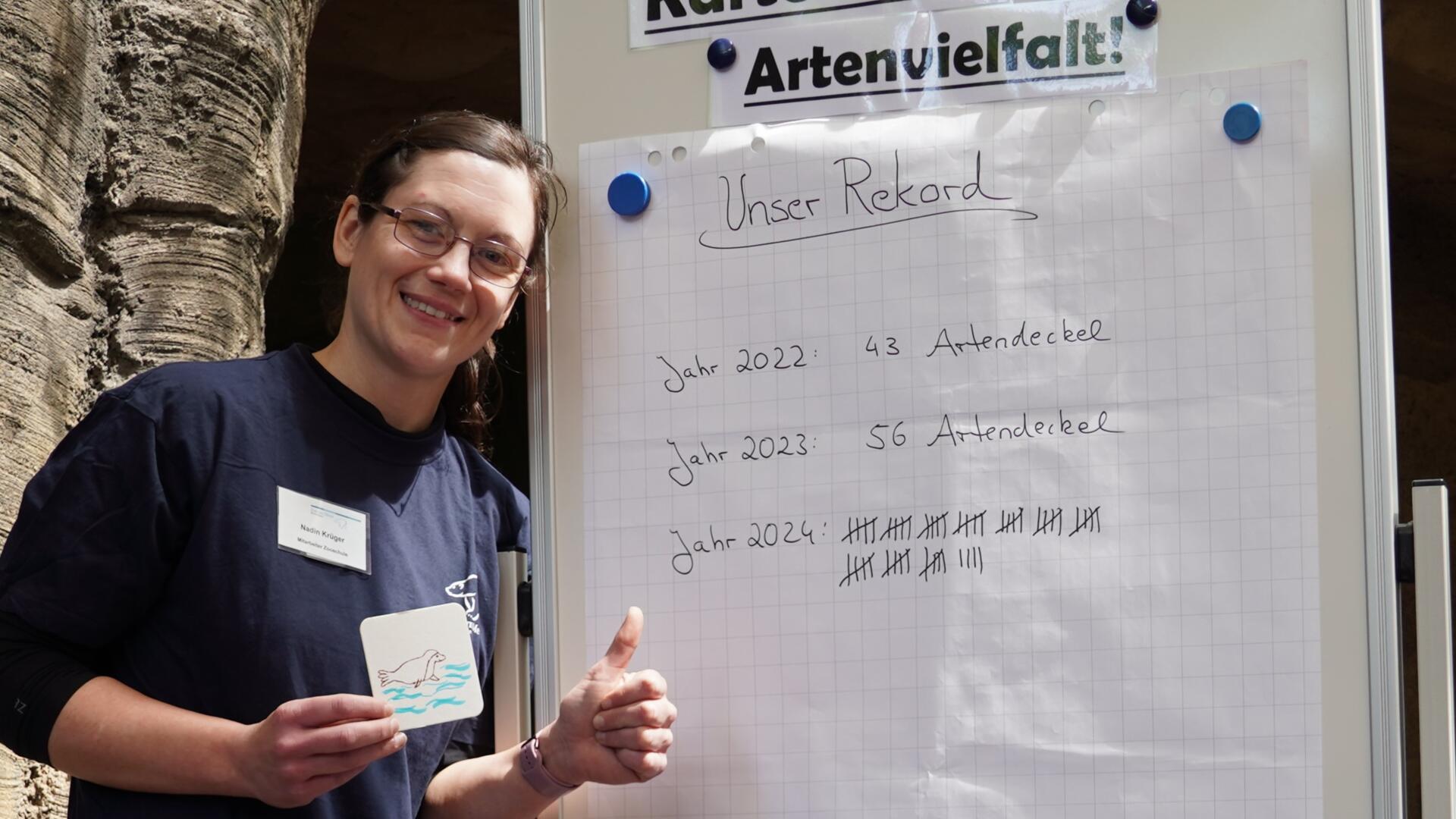 Eine Mitarbeiterin des Zoos am Meer steht lächelnd vor einer Tafel mit dem Titel „Unser Rekord“ und zeigt auf eine Liste gesammelter „Artendeckel“ der Jahre 2022 bis 2024.