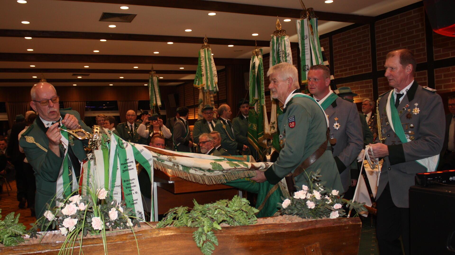 Mehrere Männer in grünen Uniformen stehen in einem Saal und präsentieren feierlich eine Fahne mit grünen Bändern. Einer von ihnen befestigt ein weiteres Band daran, während ein anderer die Fahne hält. Im Hintergrund sind weitere Uniformierte und Gäste in einem festlich dekorierten Raum zu sehen.