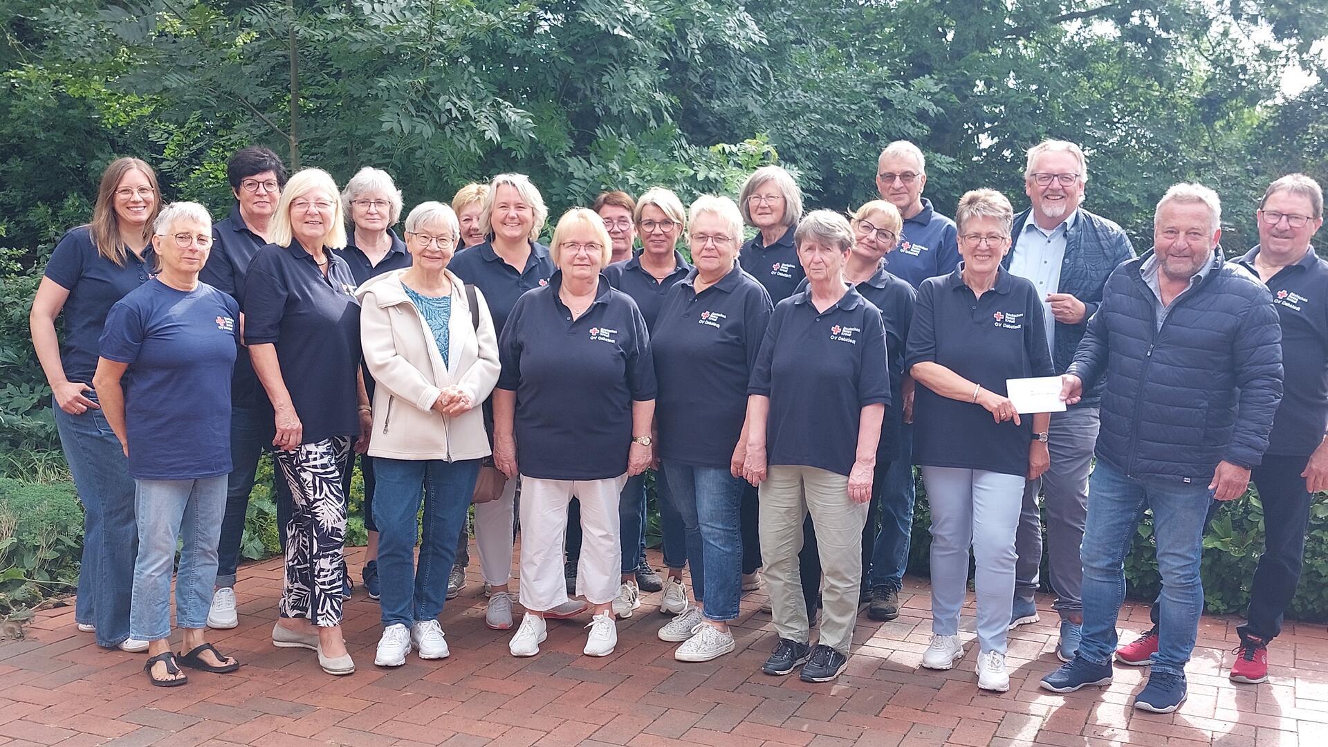 Gruppenfoto von rund 20 Mitgliedern des DRK-Ortsvereins Debstedt, die teils in dunklen DRK-Poloshirts gekleidet sind und im Freien auf einem gepflasterten Platz vor grünem Hintergrund stehen. Zwei Personen vorne rechts überreichen einen Umschlag.