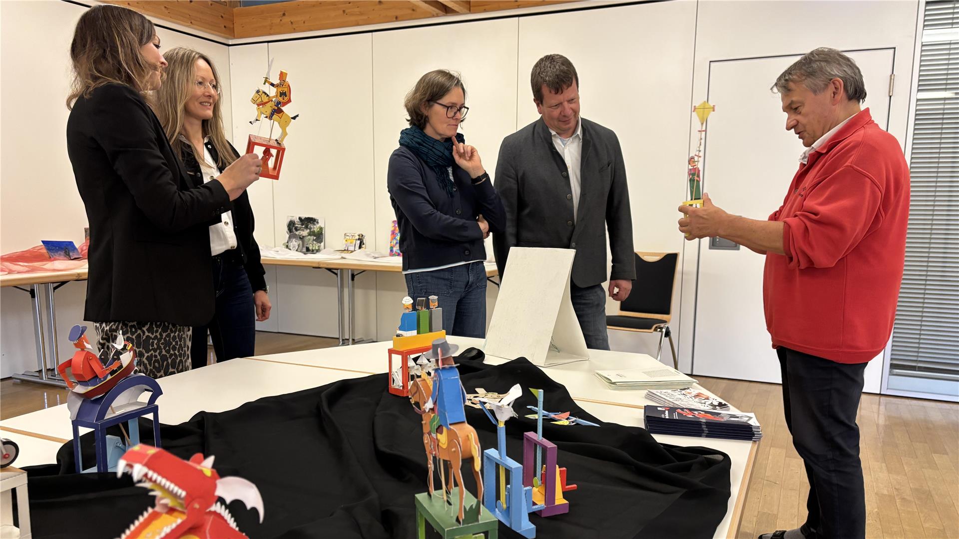 Papierkünstler Walter Ruffler zeigt mehreren interessierten Erwachsenen bunte mechanische Papierfiguren auf einer Tischpräsentation in einem Ausstellungsraum.