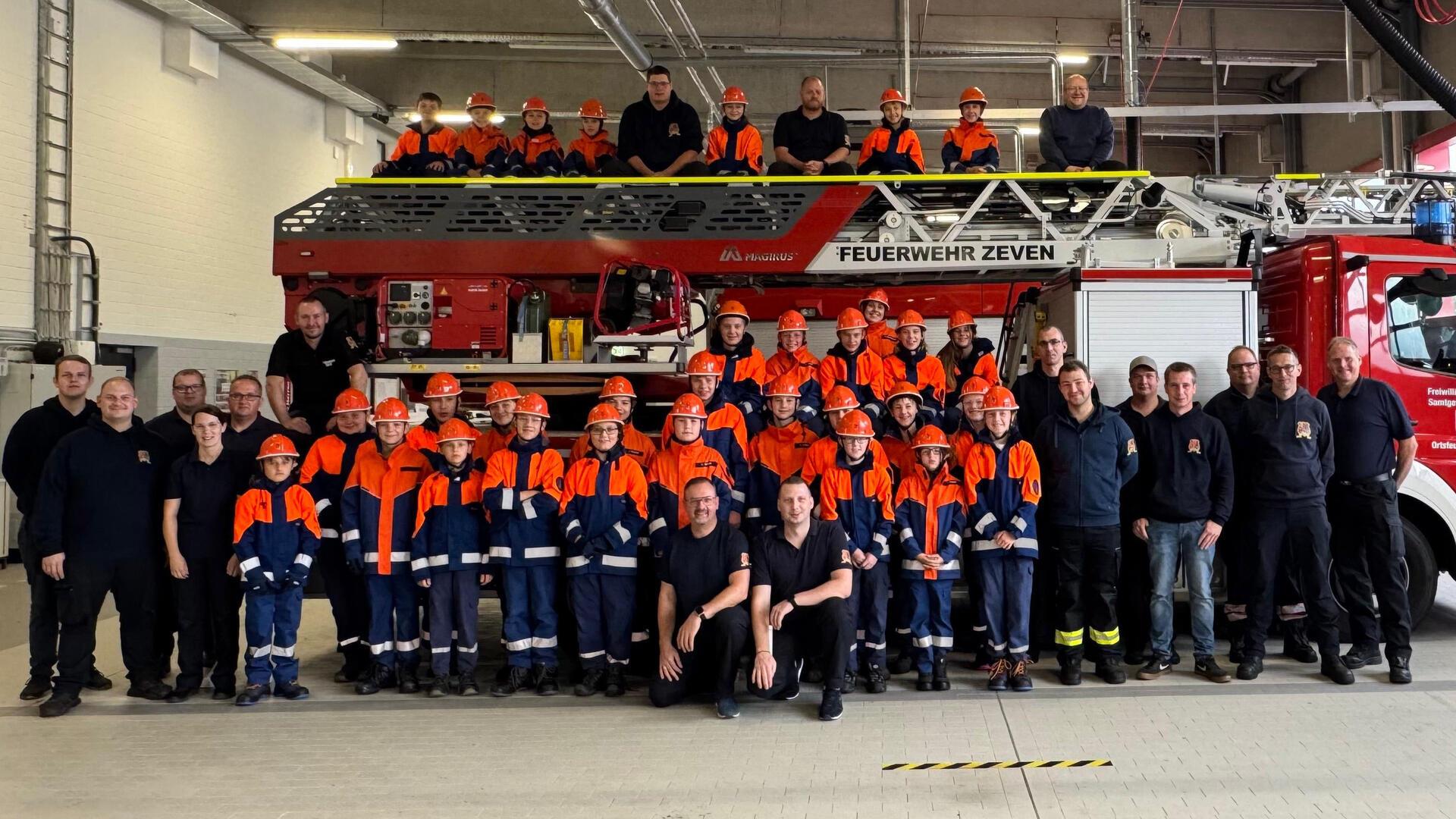 großes rotes Feuerwehrauto  und davor in blau-orangen Anzügen jugendliche Feuerwehrleute