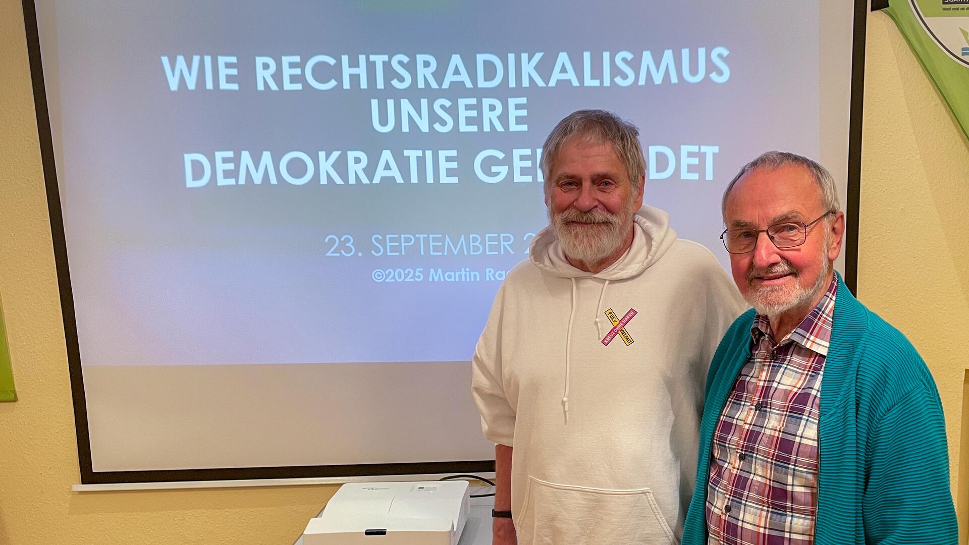 Zwei Männer stehen lächelnd vor einer Leinwand, auf der die Präsentation „Wie Rechtsradikalismus unsere Demokratie gefährdet“ angezeigt wird. Einer trägt einen weißen Kapuzenpullover mit einem Aufnäher „Kreuz ohne Haken“, der andere ein kariertes Hemd und eine türkisfarbene Strickjacke. Vor ihnen steht ein Beamer.