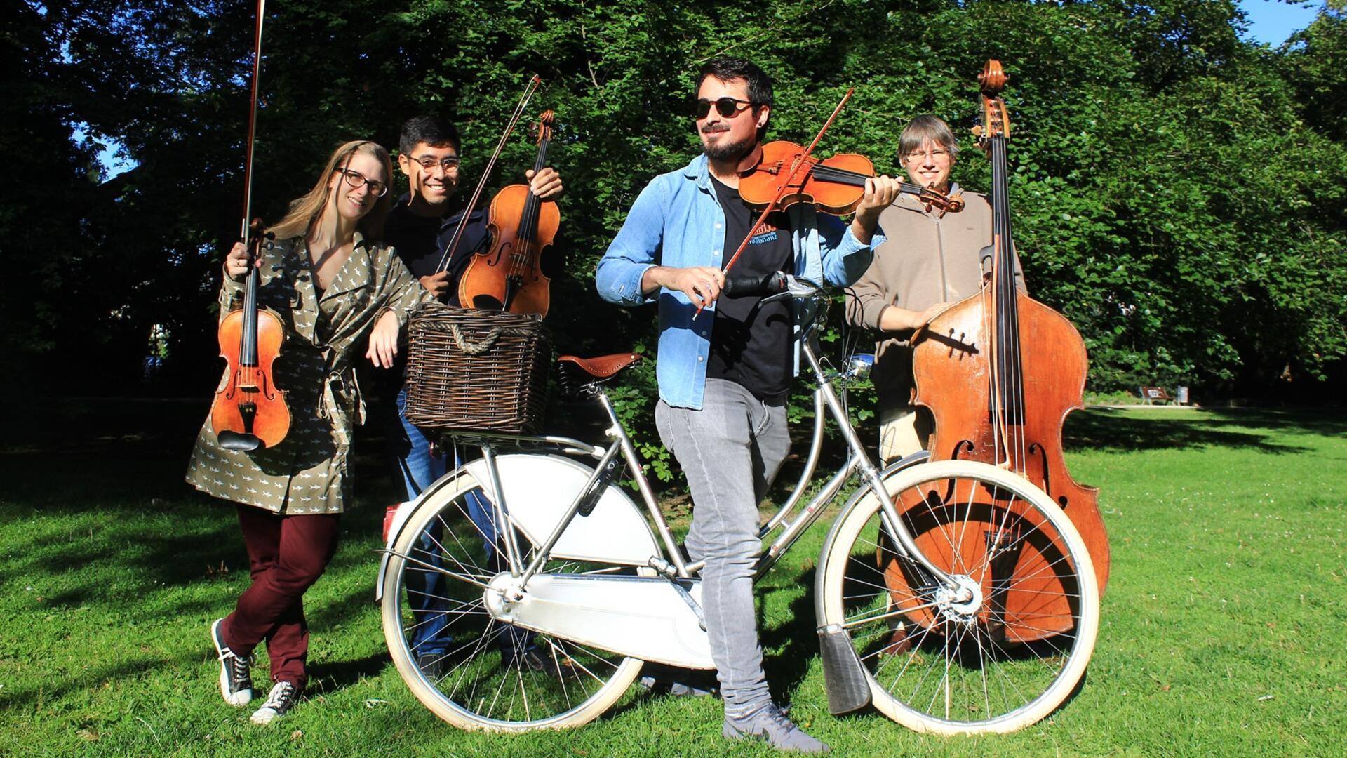 Vier Musiker stehen auf einer einer Wiese. Einer sitzt auf einem Fahrrad. Alle haben Instrumente dabei. 