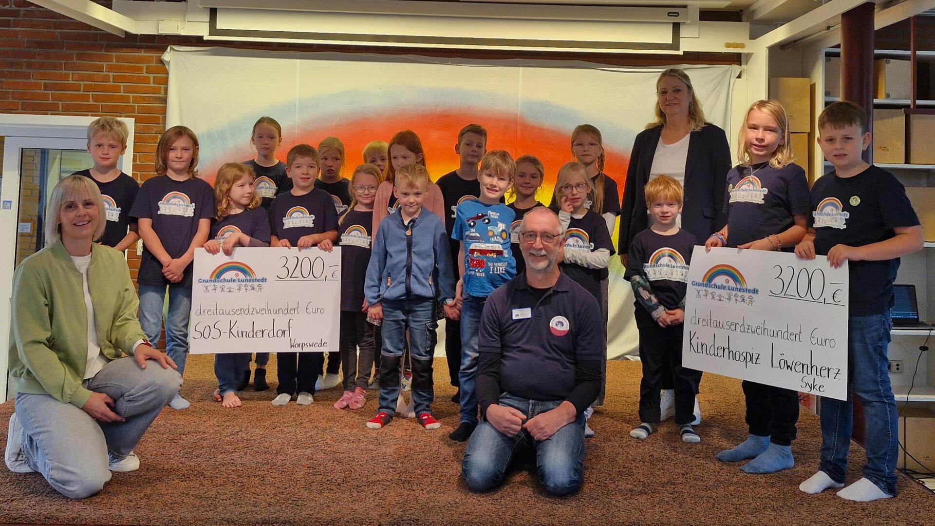 Eine Gruppe von Grundschulkindern aus Lunestedt steht gemeinsam mit zwei Erwachsenen vor einem bunten Hintergrund. Zwei große Spendenschecks über je 3200 Euro für das SOS-Kinderdorf und das Kinderhospiz Löwenherz werden präsentiert.